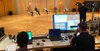 Blick über die Schultern von zwei Technikern am Mischpult auf die Bühne: Ein Kameramann filmt eine Podiumsdiskussion