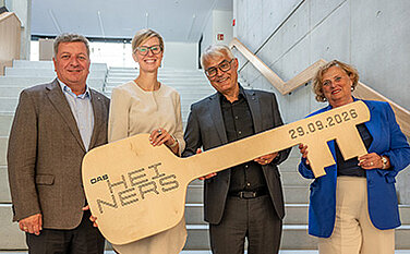 Foto von der symbolischen Schlüsselübergabe: Vier Personen halten einen großen Holzschlüssel mit der Aufschrift "HEINERS"